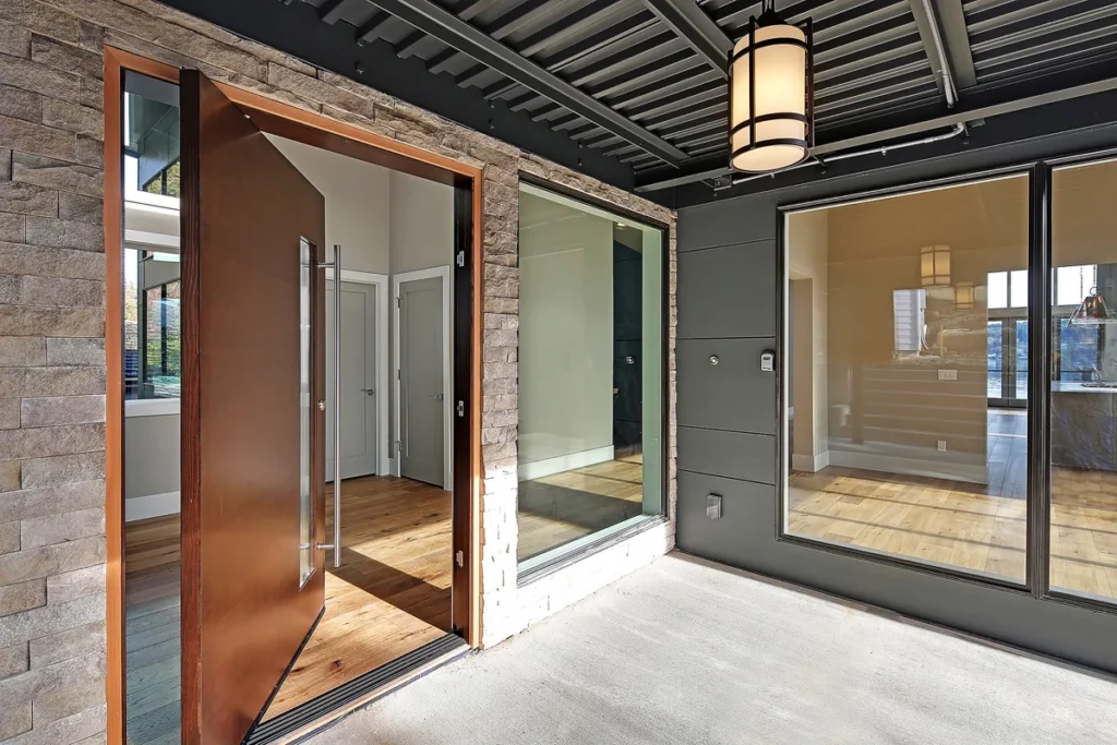Modern home entry foyer with a wood pivot door, stone accent wall, and glass surround, designed by Piper Cole Architects in the Seattle area.