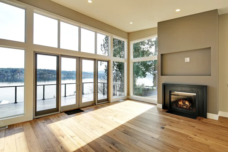Open-plan living room with a linear fireplace, wide-plank floors, and floor-to-ceiling windows framing a waterfront view, renovated by Piper Cole Architects near Seattle.