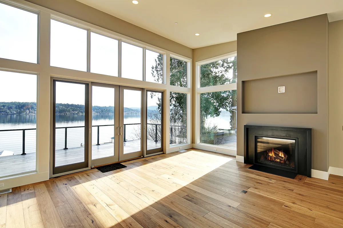 Open-plan living room with a linear fireplace, wide-plank floors, and floor-to-ceiling windows framing a waterfront view, renovated by Piper Cole Architects near Seattle.