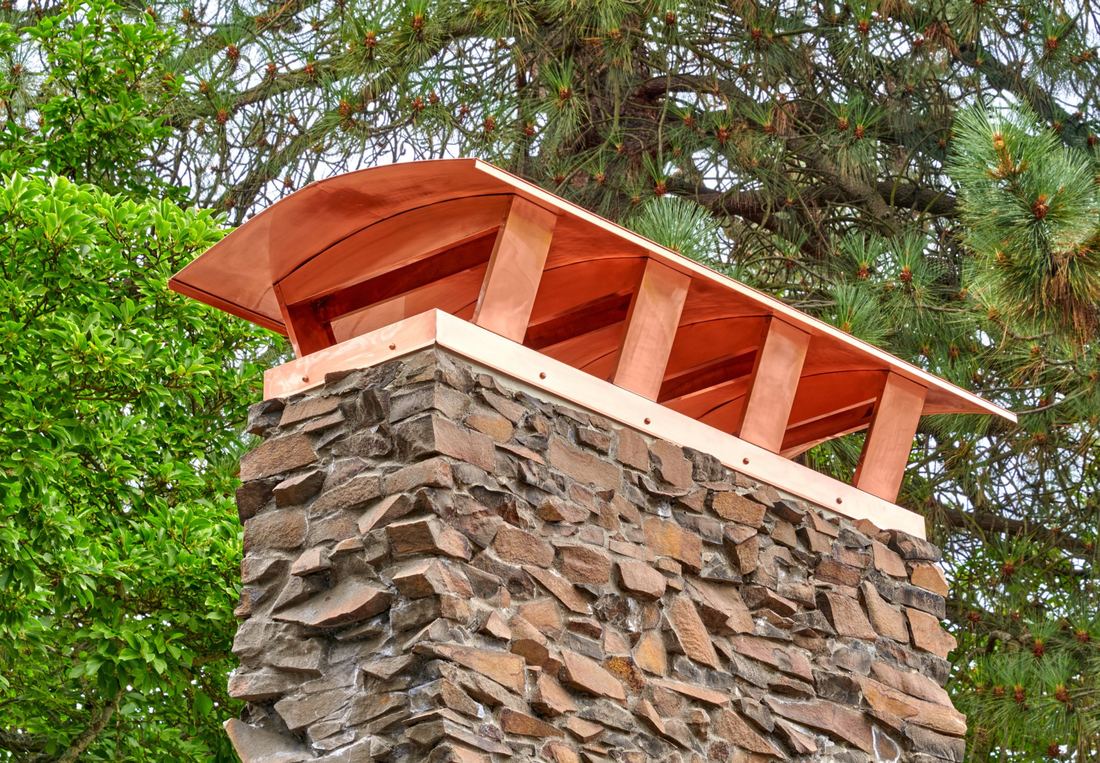 Copper chimney cap on a stacked stone chimney framed by pine branches