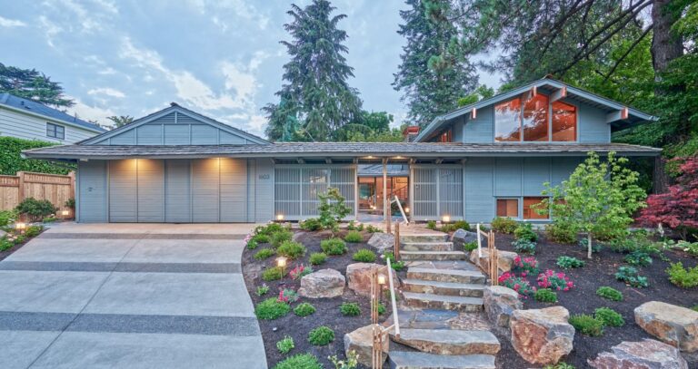 Gray midcentury house at dusk with lit entry, stone steps, boulders and new landscaping