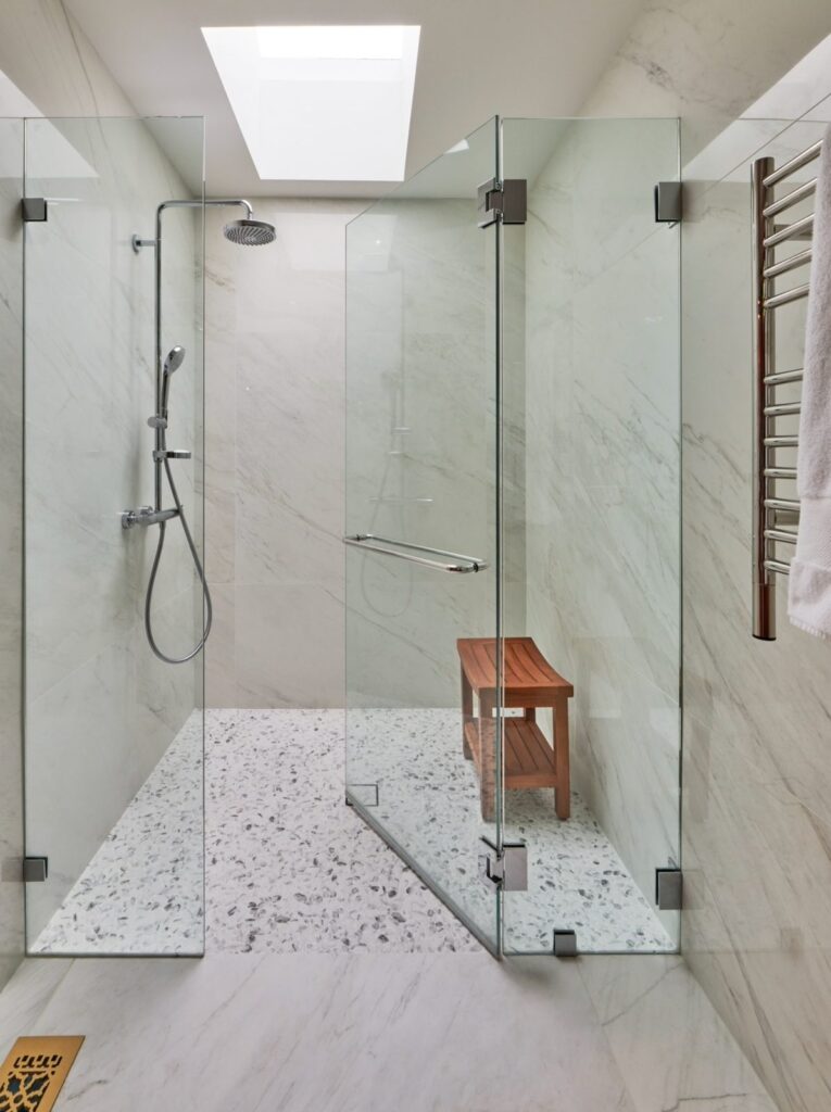Frameless glass walk-in shower with marble walls, pebble floor, teak bench and skylight