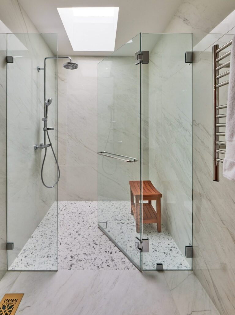 Frameless glass walk-in shower with marble walls, pebble floor, teak bench and skylight